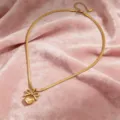 Primo piano dettagliato della collana dorata con pendente a fiocco e cuore su tessuto rosa. - Collana / Oro