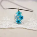 Closeup detail of an oval resin dried flower pendant necklace featuring vibrant blue pressed blooms. - Blue
