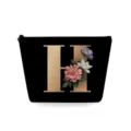Close-up van zwart cosmetisch tasje met gouden bloemletter H en gladde ritsdetails. - Hoofdletter H Stijl