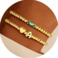 Set di due bracciali dorati con perline, cristallo verde, cuore e ciondolo lettera A. - Lettera A / Maggio