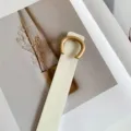 Close-up detail shot of a chic beige thin leather belt with a unique gold C-shaped buckle. - White