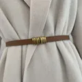 Close-up of thin brown leather belt with retro brass buckle worn over beige coat detail. - Camel