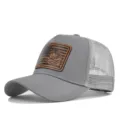 Nahaufnahme der grauen Mesh-Trucker-Mütze mit braunem Tiger-Lederaufnäher vorne. - Hellgrau