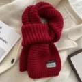 Close-up detail of a cozy red chunky knit scarf with brand label on a soft white coat background. - Red