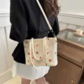 Woven straw tote bag detail showing delicate red cherry embroidery and white leather handles. - White
