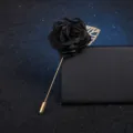 Detailed close-up of a black fabric flower brooch with a gold long pin and leaf accent. - Black