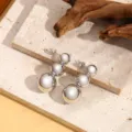 Detail van metalen hangende oorbellen met drie bollen op een natuurlijke stenen achtergrond. - A002 Stijl / Witgoud