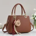 Braune Lederhandtasche Detail mit süßem Plüschbären-Anhänger und elegantem Herz-Metallanhänger. - Erdgelb