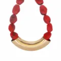 Detailed close-up view of the red resin bead necklace featuring a bold curved gold metal tube pendant. - Red