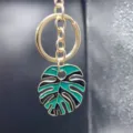 Detailed close-up of a stylish gold metal keychain featuring a green and black monstera leaf pendant. - Style T