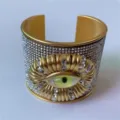 Brede gouden kleurige manchetarmband met fonkelende strass steentjes en surrealistisch oog. - Stijl A (Goud)