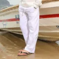 Detailed view of men's casual white cotton linen trousers with drawstring waist on beach. - S / Bottoms / White