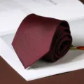 Detailed view of a burgundy textured necktie rolled neatly on a white book for business wear. - Hand-Tied / 8cm / Calm Burgundy