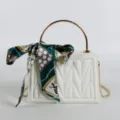 White structured crossbody bag with gold metal handle and stylish printed silk scarf accessory. - White