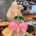 Close-up view of a stylish pink crystal cherry keychain and bag charm with gold metal details. - Pink Iridescent Green Leaf Cherry