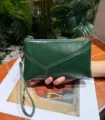 Detailed shot of dark green leather wristlet clutch bag, showcasing fine texture and wrist strap. - Green
