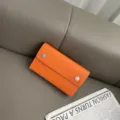 Detailansicht der orangefarbenen Leder Umhängetasche mit Druckknöpfen auf grauem Sofa. - 19*11.5*4.5CM / Orange