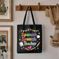 Detail of black canvas tote bag with colorful Spanish teacher print hanging on a wooden wall hook. - Style 1 / Black