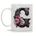 Close-up of white ceramic coffee mug featuring gothic letter G and pink rose floral design. - Letter G