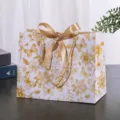 Witte papieren geschenkzak met gouden bloemenprint en elegant striklint voor feestelijke verpakking. - Kleine Maat 18x16x10cm / Goud Bloemen met Tekst (Gouden Lint)