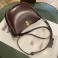 Detailed view of the dark burgundy leather saddle bag with gold metal lock and shoulder strap. - Coffee
