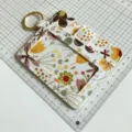 Detailed closeup of a floral print ID card holder with golden keyring on a grid craft mat. - Khaki