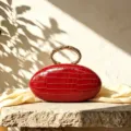 Detailed view of red PU crocodile texture oval clutch bag featuring elegant gold metal ring handle. - Red
