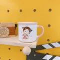Close-up detail of a cute cartoon girl ceramic mug displayed on a wooden shelf background. - 301-400ml / Pink