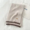 Primer plano de una cómoda bufanda de punto beige con bordes marrones contrastantes y etiqueta. - Beige