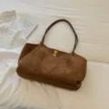 Vista detallada del bolso tote de ante marrón con asas finas y cierre dorado en cama blanca. - Marrón