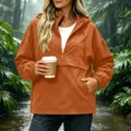 Vue detaillee de la veste impermeable a capuche orange pour femme dans une foret pluvieuse. - S / Hauts / Orange