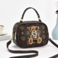 Vista dettagliata della borsa a tracolla marrone con monogramma, toppa d'orso e metalleria. - Caffè