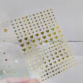 Detailed view of 3D gold foil mini heart nail stickers with adhesive backing on clear sheet. - DH-539 / Gold