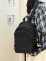 Minimalist black backpack detail shot, showing fabric texture, front pocket, and cute cat pendant. - With Charm / Black