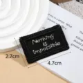Detail view of a black embroidered clothing patch with white text and size dimensions. - Nothing / Black