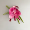 Detailed view of artificial pink orchid boutonniere with green foliage and red ribbon decor. - Large Phalaenopsis Boutonniere / L