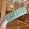 Close-up detail of sage green leather square hair clip with embossed love heart pattern. - Green