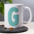 Close-up of a white ceramic coffee mug featuring a bold teal letter G design on slate. - 19