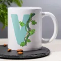 Close-up detail of white ceramic coffee mug with letter V and green vine leaf pattern print. - 22