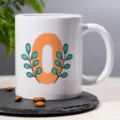 White ceramic coffee mug with stylish orange letter and teal leaf design for office desk. - 24