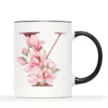 Ceramic coffee mug detail view featuring elegant pink floral monogram design and black handle. - Letter V