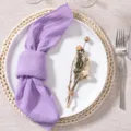 Close-up view of a purple wrinkled fabric napkin styled on a white plate with dry flower. - 32*32 / Purple