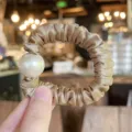 Detail shot of elegant champagne satin hair scrunchie adorned with a large faux pearl. - M / Brown