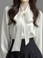 Close-up detail of elegant white satin bow tie blouse with long sleeves and pleated skirt. - White