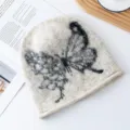 Nahaufnahme der flauschigen Strickmütze mit schwarzem Schmetterlingsmuster für kalte Tage. - L (56-62cm) / Gebrochenes Weiß