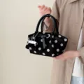 Black polka dot handbag with twin handles and a silver charm, shown held by a model - Black