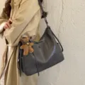 Gray faux-leather shoulder bag with teddy charm, shown on a model in a beige trench coat - With Pendant / Gray