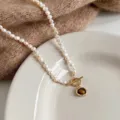 Primo piano di perle bianche irregolari, chiusura dorata e ciondolo in pietra marrone su un piatto - Collana con Ciondolo Occhio di Tigre / 45cm / Bianco