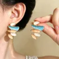 Primo piano di orecchini smaltati irregolari blu, crema e pesca, indossati e tenuti in mano - Orecchini a Bottone Smalto Astratto (0056) / Oro