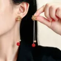 Primo piano di lunghi orecchini dorati con catenelle e sfere rosse, indossati all’orecchio - Stile Oro 1 (1956)